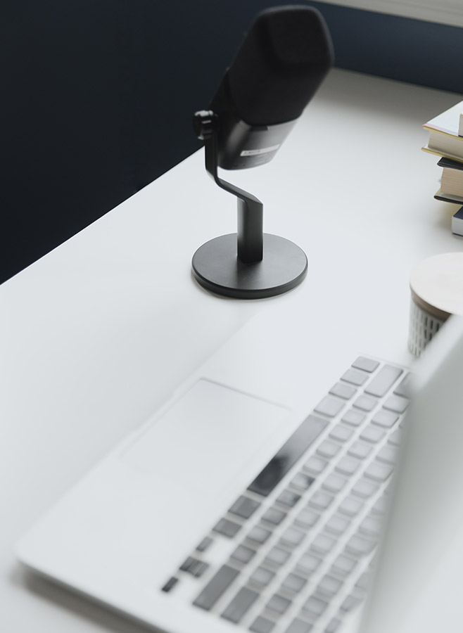 A close-up of a modern desk with a laptop, a black microphone on a stand, a closed notebook, and a stack of books in the background—perfect for hosting virtual mental health workshops in a clean, minimalistic workspace.