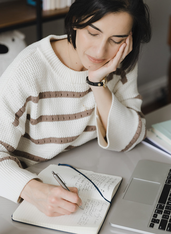 A trauma psychologist with dark hair, wearing a white and brown striped sweater, sits at a desk, resting her head on one hand while writing in a notebook next to a laptop. She appears focused and relaxed.
